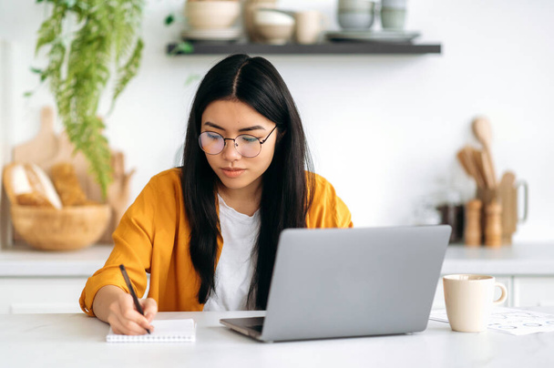 Онлайн-образование, концепция электронного обучения. Умная позитивная азиатская девушка в очках, успешная студентка, учится из дома с помощью ноутбука, слушает онлайн-вебинар, уроки, делает заметки, улыбается дружелюбно - Фото, изображение