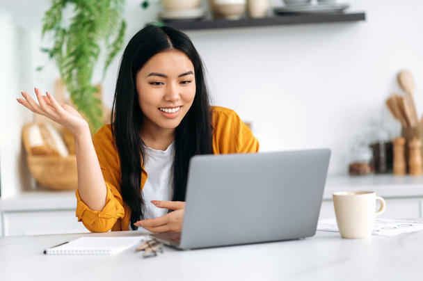 Онлайн відеозв'язок. Дружня азіатська брюнетка молода жінка, менеджер, фрілансер, сидить на кухні, розмовляючи на відеодзвінок ноутбуком з клієнтом або співробітниками, обговорюючи ідеї, посміхаючись
 - Фото, зображення