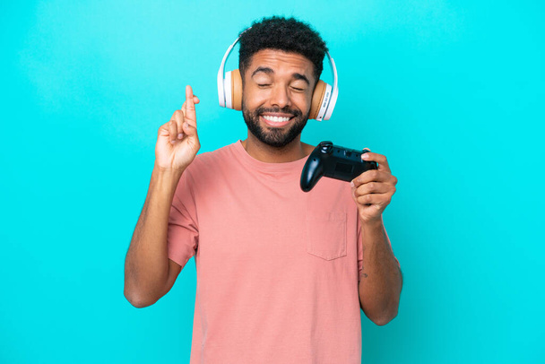 Young brazilian man playing with a video game controller isolated on blue background with fingers crossing and wishing the best - Zdjęcie, obraz