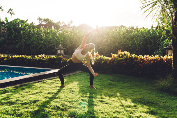 Blond woman in sportswear and barefoot doing yoga practice while standing in Revolved Chair with Arms Extended pose on green lawn - Photo, image