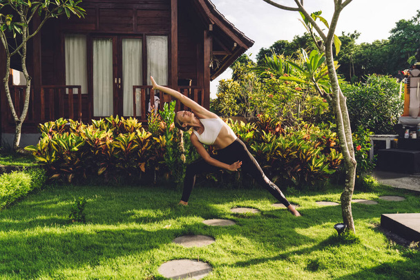 Concentrated strong female wearing sportswear doing yoga in Extended Side Angle pose while spending time in tropical garden in summer - Φωτογραφία, εικόνα