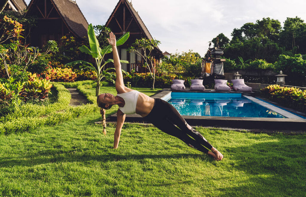 Slim woman practicing yoga exercise and doing stretches while living healthy lifestyle and enjoying training alone in green countryside area near pool - Foto, immagini