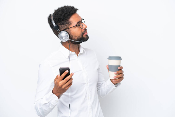 Telemarketer Βραζιλίας άνθρωπος που εργάζονται με ένα ακουστικό απομονώνονται σε λευκό φόντο κρατώντας καφέ για να πάρει μακριά και ένα κινητό - Φωτογραφία, εικόνα