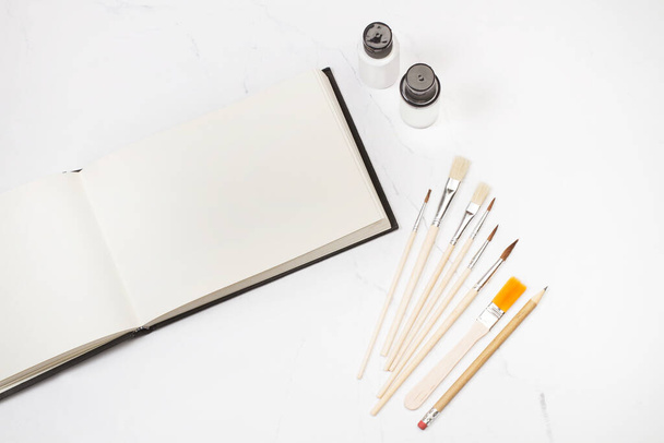 Wooden artistic paintbrushes, paint bottles and a paper notebook on a white marble - Fotoğraf, Görsel