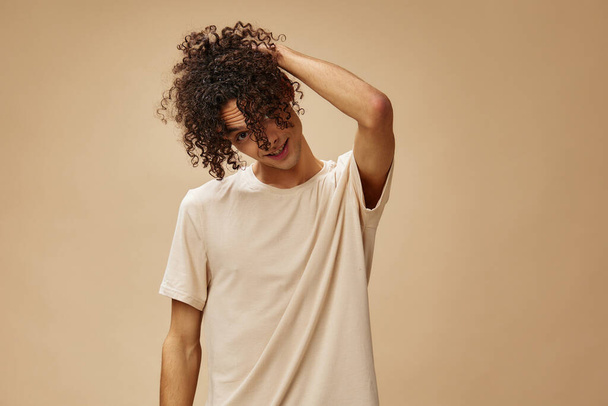 Funny awesome tanned curly man in basic t-shirt touches hair curls posing isolated on over beige pastel background. Fashion New Collection offer. People and Emotions concept. Free place for ad - Photo, Image