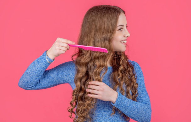 vrolijk tiener meisje kammen krullend haar met haarborstel op roze achtergrond. - Foto, afbeelding