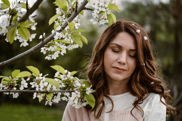 Beautiful adult woman closed eyes, enjoying flowers and smelling blossom tree in the spring garden. Close-up portrait with natural makeup - Foto, Imagen