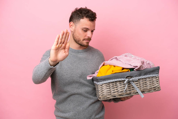 Young Brazilian man holding a clothes basket isolated on pink background making stop gesture and disappointed - Photo, Image