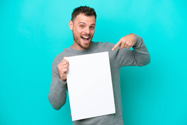 Fiatal brazil férfi, kezében egy üres plakáttal, kék háttérrel, kezében egy üres plakáttal, boldog arckifejezéssel és rámutatva. - Fotó, kép