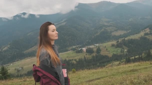 Female walks green grass against backdrop beautiful mountains covered with white clouds. Magic view, mountain village, side view. Concept of travel, go to the goal, confident, strong, independent - Footage, Video