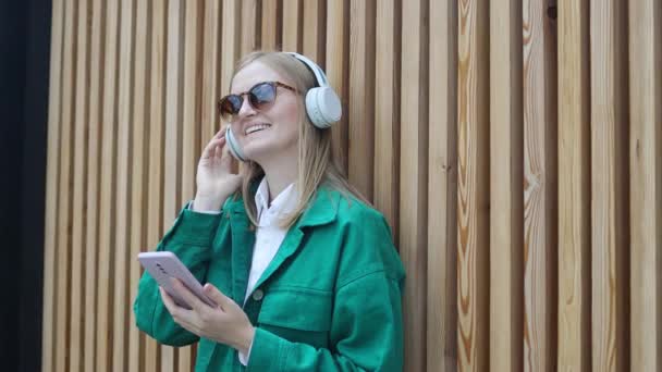 Funny young blonde 30s woman in casual green jacket eyeglasses on wood background. People lifestyle concept. Listen music with air pods hold mobile phone, dancing.  - Video