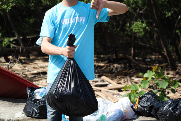 Ehrenamtliche im blauen T-Shirt mit Müllsack und Daumen nach unten auf Müllkippe, Ökologie-Mann säubert und sammelt Müll auf, um Umweltverschmutzung zu reduzieren Umweltproblem, Weltumwelttag. - Foto, Bild