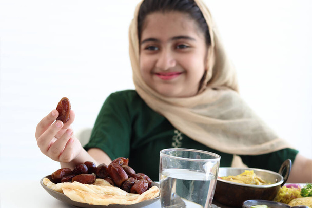 Adorable smiling Pakistani Muslim girl with beautiful eyes with hijab, hold date fruits (sweet and high energy food mostly eat in Ramadan of Islam), enjoy eating Islamic halal food on white background - Photo, image