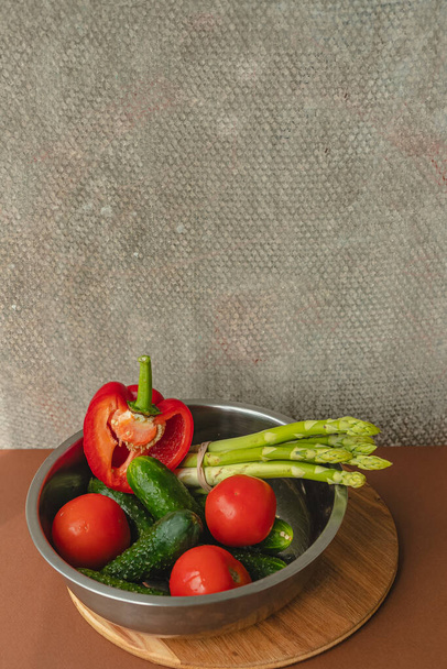 Τα λαχανικά βρίσκονται σε ένα μεταλλικό μπολ: ντομάτες, σπαράγγια, αγγούρια, κόκκινες πιπεριές. σε ξύλινη σανίδα και καφέ φόντο. πίσω γκρι φόντο. τόπος για κείμενο. - Φωτογραφία, εικόνα