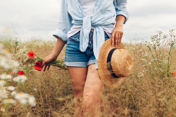 Modaya uygun kırsal yaz aksesuarları ve giysileri. Hasır şapkalı kadın çayırda keten gömlek giyip kır çiçeklerini topluyor.. - Fotoğraf, Görsel