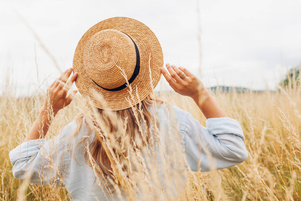 Female fashion. Back view of young woman walking among high grasses in summer meadow wearing straw hat and linen shirt. Stylish accessories and clothing - Фото, изображение