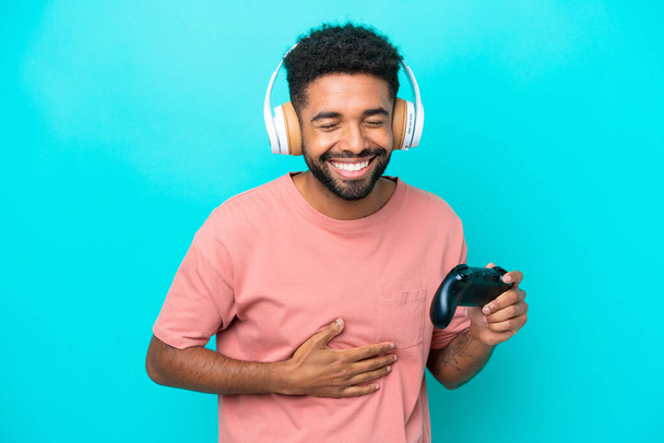 Young brazilian man playing with a video game controller isolated on blue background smiling a lot - Foto, Imagem