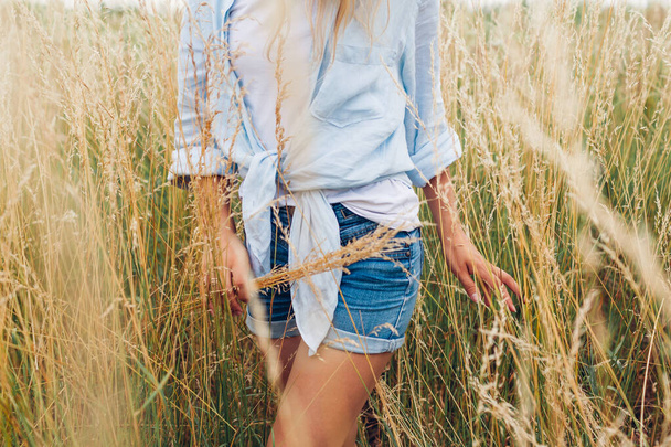 Çayırda yürüyen, keten gömlek ve kot gömlek giyen çimen toplayan genç bir kadın. Doğal yaz kıyafetleri. Konforlu kumaş - Fotoğraf, Görsel