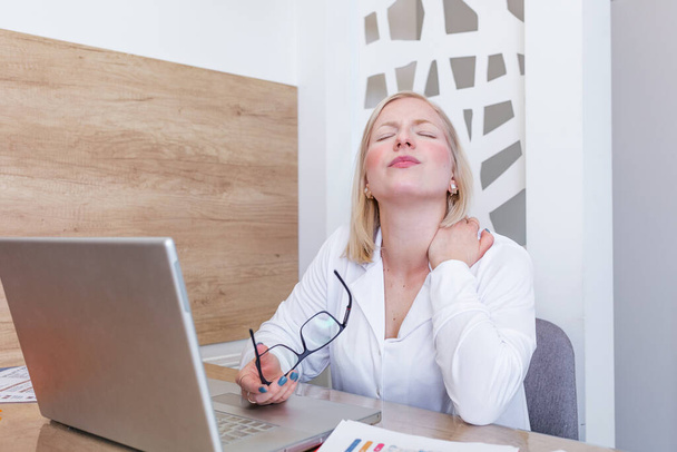Geschäftsfrau verspürt Nackenschmerzen, nachdem sie mit Laptop am Tisch sitzt. Müde Frauen leiden am Bürosyndrom, weil sie stundenlang am Computer arbeiten. hübsches Mädchen massiert ihre angespannten Nackenmuskeln - Foto, Bild