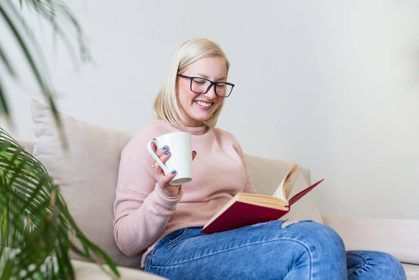 Young woman at home sitting on modern sofa relaxing in her living room reading book and drinking coffee or tea. White cozy bed and a beautiful girl, reading a book, concepts of home and comfort - Foto, Imagem