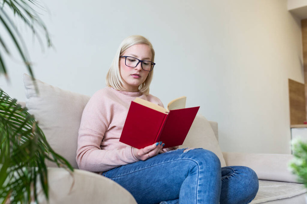 Young beautiful woman lying on sofa and reading book at home. Excellent book. Beautiful young woman reading a book at the sunny morning. - Foto, Imagem