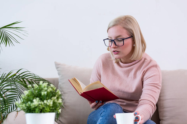 Genç bir kadın modern koltukta oturmuş oturma odasında kitap okuyor ve kahve ya da çay içiyor. Beyaz rahat bir yatak ve güzel bir kız, kitap okuyor, ev kavramları ve rahatlık. - Fotoğraf, Görsel