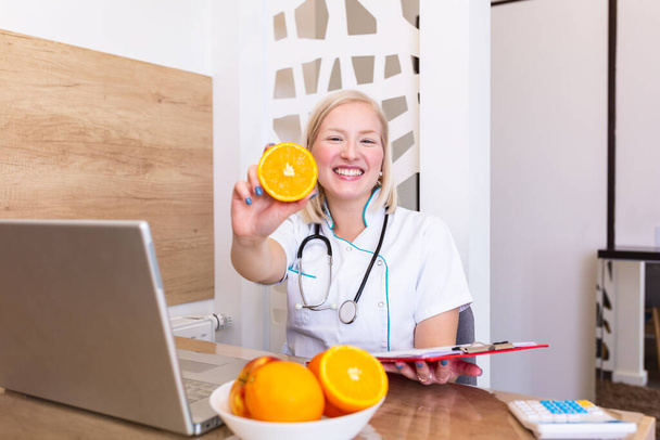 Retrato de una joven nutricionista sonriente en la sala de consulta. Escritorio nutricionista con fruta sana, jugo y cinta métrica. Dietista trabajando en plan de dieta
. - Foto, Imagen