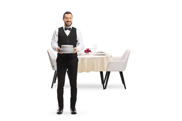 Full length portrait of a server holding a pile of plates and standing in front of a table isolated on white background - Fotó, kép