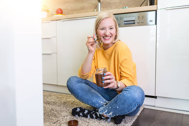 Nette Albino junge Frau in modernen stilvollen Kleidern genießen leckeren Schokoladenaufstrich mit süßem Lächeln in der Küche Interieur. junge Frau isst Schokolade aus einem Glas, während sie auf dem Küchenboden sitzt. - Foto, Bild