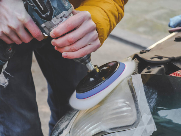 The hands of a young caucasian guy in a yellow jacket holds a drill with a microfiber disc and polishes the front headlight of his car on a city street, close-up side view. Home renovation concept. - Photo, Image