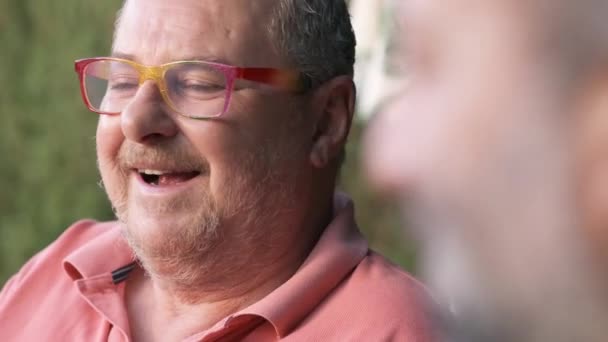 Content middle aged male in eyeglasses looking at crop anonymous friend while sitting on street against blurred background on summer day - Séquence, vidéo