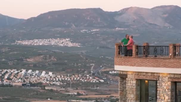Mężczyźni w jasnych koszulkach całujący się podczas randki na tarasie kamiennego budynku przed górami o zachodzie słońca - Materiał filmowy, wideo