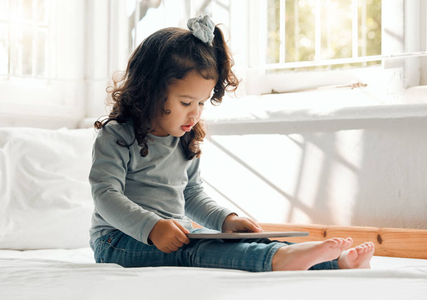 Shot of an adorable little girl sitting on her bed with a digital tablet. - 写真・画像