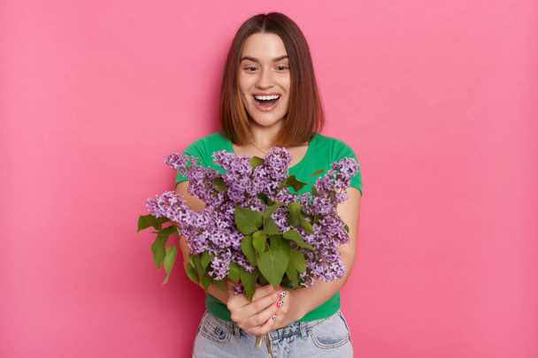 Image of excited woman standing isolated over pink background, looks with amazement at her bouquet, wearing green t shirt, carries lilac flowers, keeps mouth opened, being pleasantly surprised. - Fotografie, Obrázek
