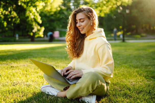Giovane donna con cuffie wireless che lavora online o studia seduta sull'erba al parco. Affari, blogging, freelance, concetto di istruzione. - Foto, immagini