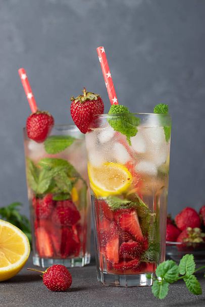 Cocktail freddo e luminoso alla fragola Mojito con menta, fette di limone, cubetti di ghiaccio e paglia da bere su sfondo grigio, formato verticale - Foto, immagini