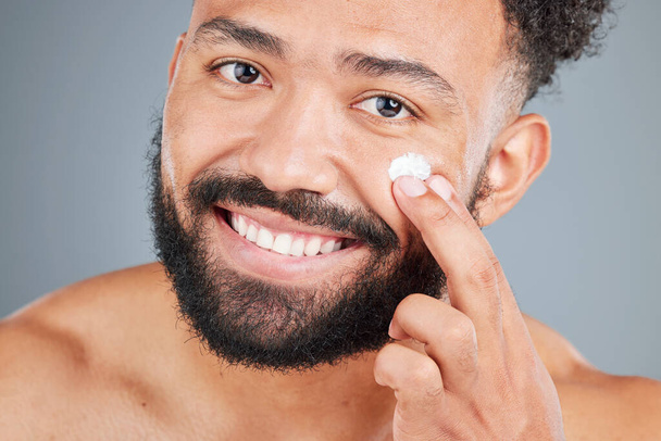 Studio portrait of a handsome young man applying lotion to his face against a grey background. - Photo, image