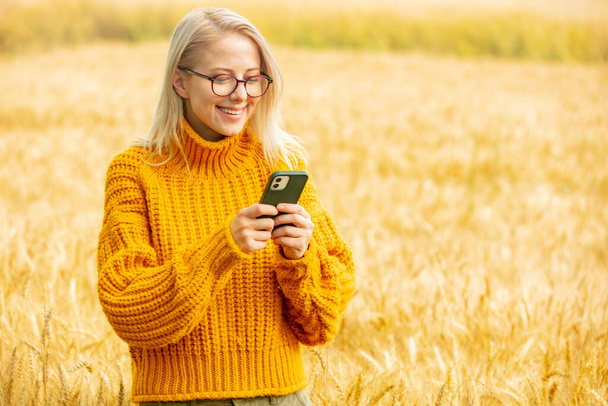 Stylisches Mädchen mit Brille und gelbem Pullover mit Handy im Weizenfeld - Foto, Bild