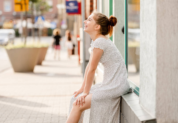 Beautiful girl in dress sitting at street in city and looking at sky in sunny day. Pretty female teenager model outdoors in summer time - Valokuva, kuva