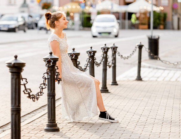 Beautiful girl in long dress sitting at chain fence at street in city and looking back. Pretty female teenager model outdoors in summer time - Foto, Imagem