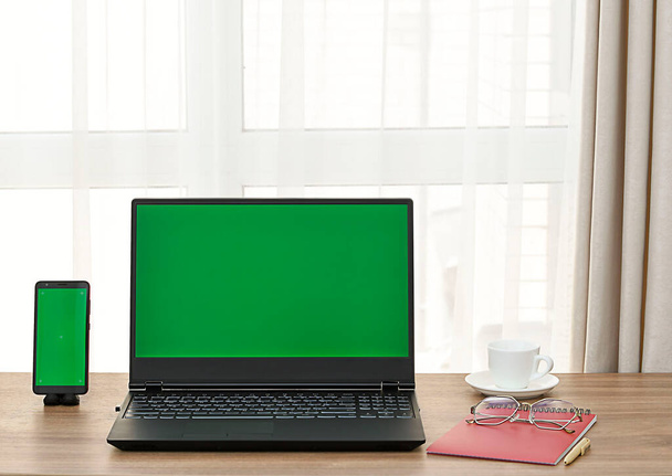 Ключ Chrome. Ноутбук и мобильный телефон с зеленым экраном на деревянном столе с чашкой кофе, блокнотом и стаканами у окна. Интерьер дома. - Фото, изображение