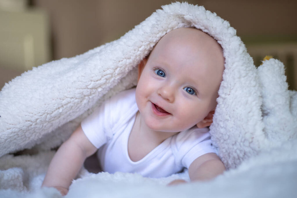 Улыбающийся малыш смотрит в камеру под белым одеялом, полотенцем. селективный фокус - Фото, изображение