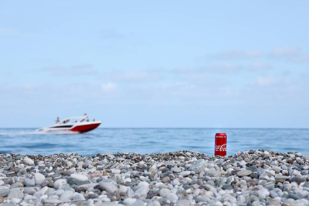 ANTALYA TURKEY - MÁJUS 18, 2021: Eredeti Coca Cola piros ón fekszik kis kerek kavicsos kövek közelében a tengerparton. Coca-kólás doboz és motorcsónak a török tengerparton - Fotó, kép