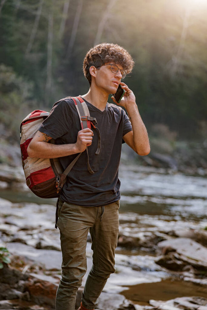 Young man tourist with backpack roaming at river in mountains and talking on cellphone. Vertical. - Foto, imagen