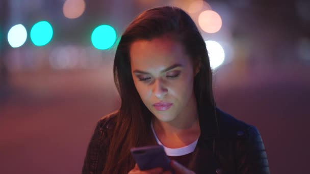 Uma jovem sorrindo e lendo textos em seu celular à noite na cidade. Uma mulher à noite usando seu smartphone. Uma jovem mulher percorrendo aplicativos em seu celular à noite. - Filmagem, Vídeo