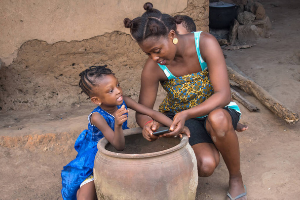  Een Afrikaanse moeder met haar kinderen buigt laag naast een grote klei pot of kalebas in de voorkant van een dorp modderhuis Een Afrikaanse moeder met haar kinderen buigen laag naast een grote klei perceel in de voorkant van een dorp modderhuis met een mobiele telefoon  - Foto, afbeelding