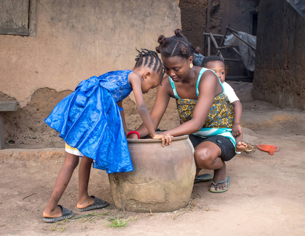  Eine afrikanische Mutter mit ihren Kindern beugt sich neben einem großen Tontopf oder Kalebasse vor einem Lehmhaus im Dorf  - Foto, Bild