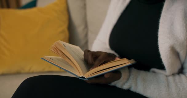 Крупный план девушки африканской внешности с книгой в синей обложке, читающей и бегущей пальцем по линии. Девушка одета в белый кардиган, сидя на диване, на котором лежат желтые и синие подушки. - Кадры, видео