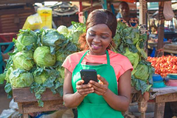 Happy African business woman or female trader wearing a green apron and using a smart phone while standing at her vegetable stall in a market place - Photo, Image
