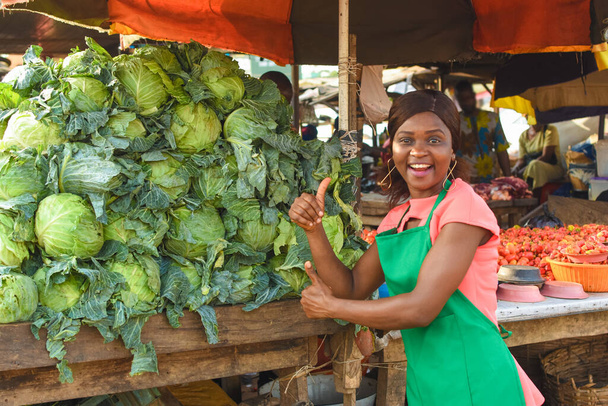 Happy African business woman or female trader wearing a green apron and doing thumbs up gestures while standing at her vegetable stall in a market place - Fotoğraf, Görsel